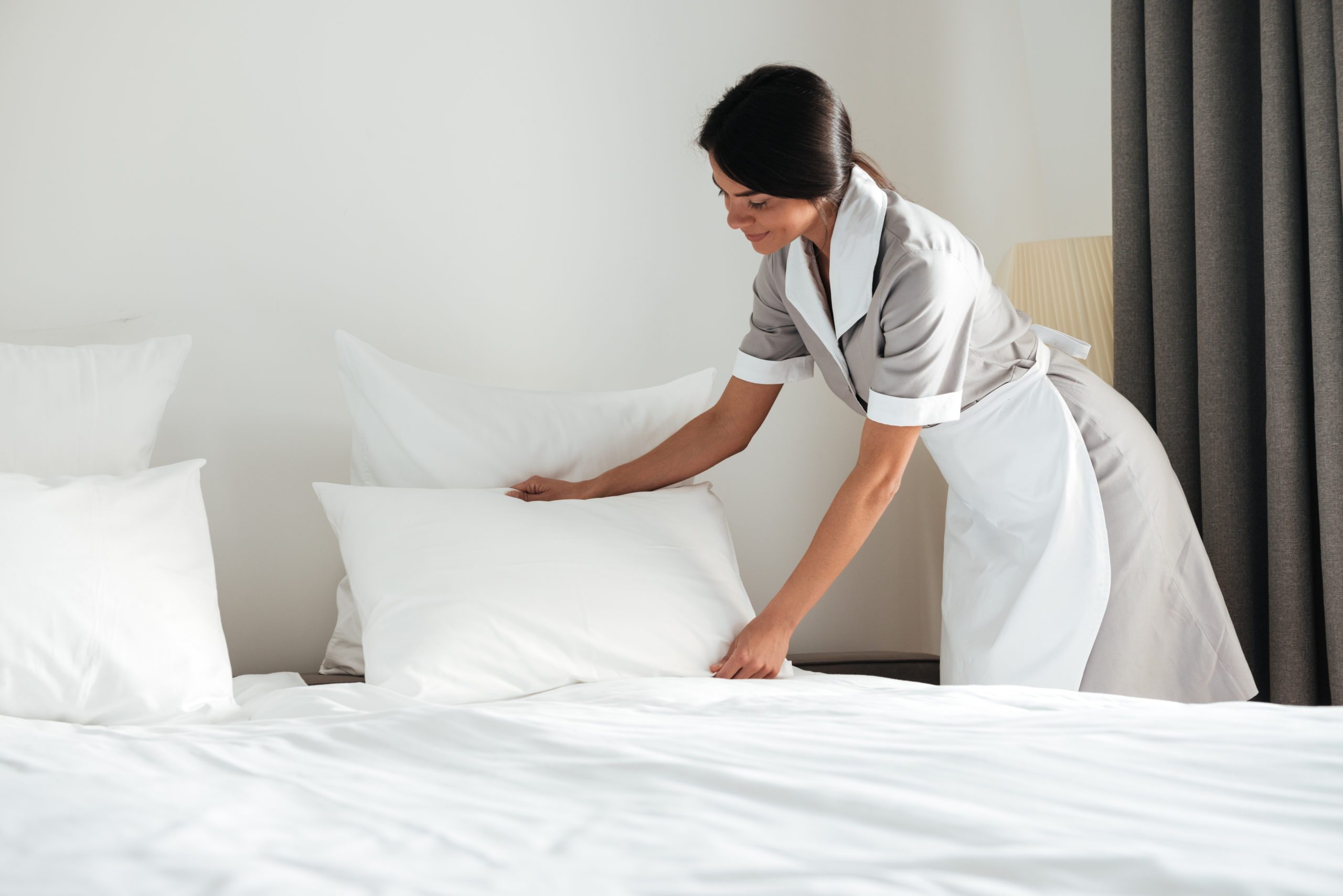 Young hotel maid setting up pillow on bed